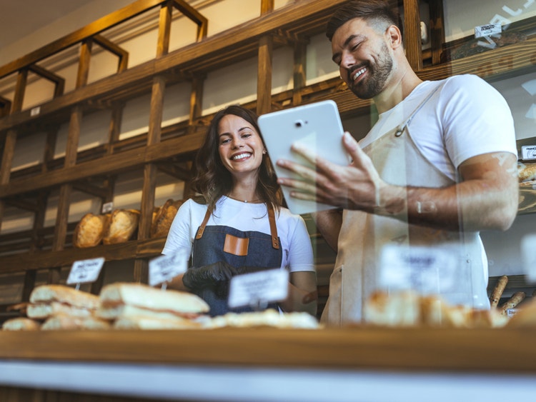 Coppia di fornai sorridenti controlla ordini su un tablet dietro il bancone pieno di pane e panini in una panetteria.