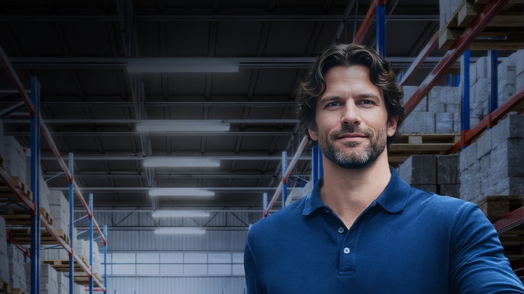 Uomo sorridente in polo blu in primo piano all&#39;interno di un grande magazzino con scaffali industriali.