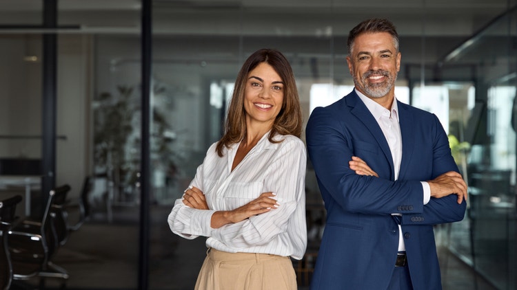 Due professionisti sorridenti, un uomo in giacca e una donna, posano a braccia conserte in un ufficio moderno.