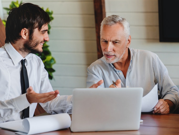 Due uomini discutono davanti a un laptop e documenti cartacei durante una consulenza professionale in ufficio.