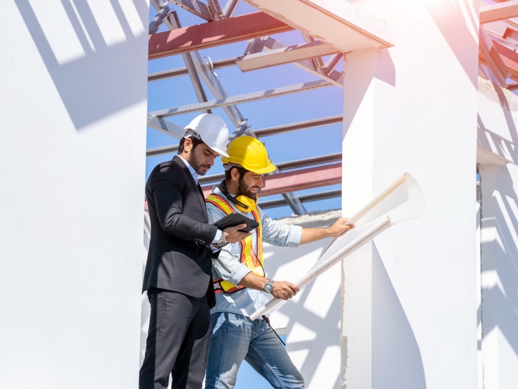 Due tecnici con casco protettivo analizzano planimetrie e tablet in un cantiere edile sotto un cielo sereno.