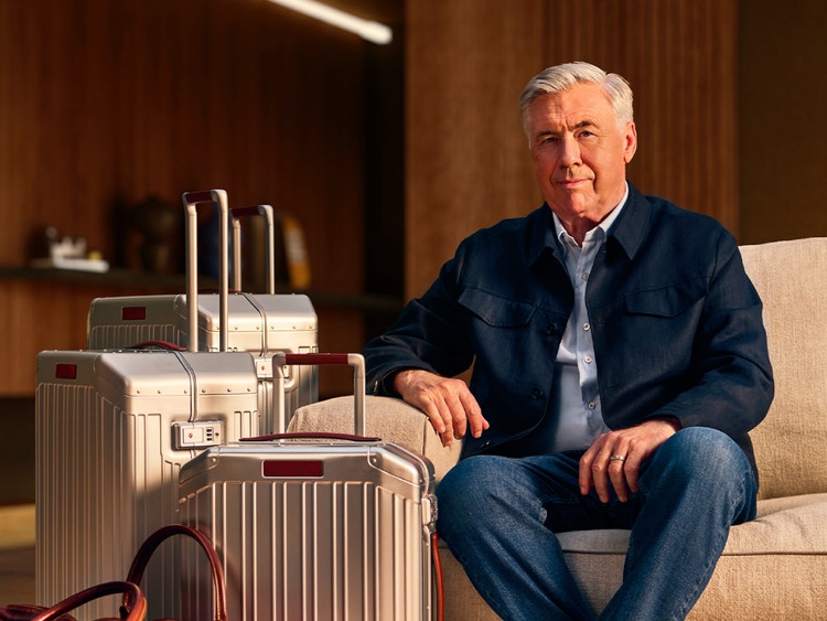Carlo Ancelotti seduto su un divano accanto a tre valigie rigide di alluminio in un ambiente elegante.