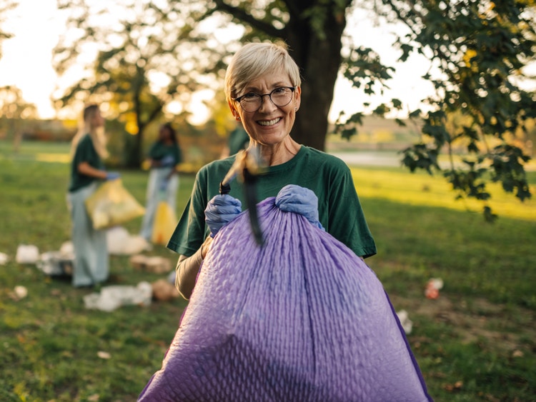 Una donna sorridente con maglietta verde tiene un grande sacco viola durante una raccolta di rifiuti in un parco.