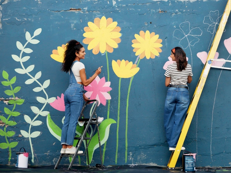Due ragazze sulle scale dipingono un grande murale colorato con fiori gialli e rosa su una parete blu scuro.