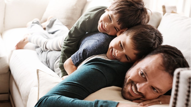 Un padre sorridente sdraiato sul divano gioca e si abbraccia con i suoi due figli piccoli.