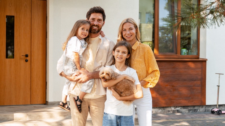 Famiglia sorridente con due figlie e un cagnolino in braccio davanti all&#39;ingresso di casa.