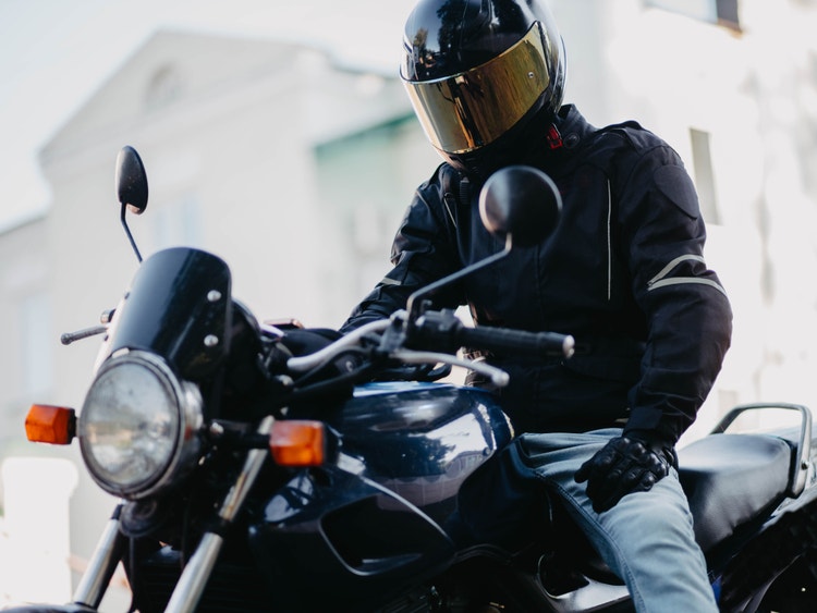 Motociclista in giacca nera e casco con visiera dorata seduto su una moto scura in strada.