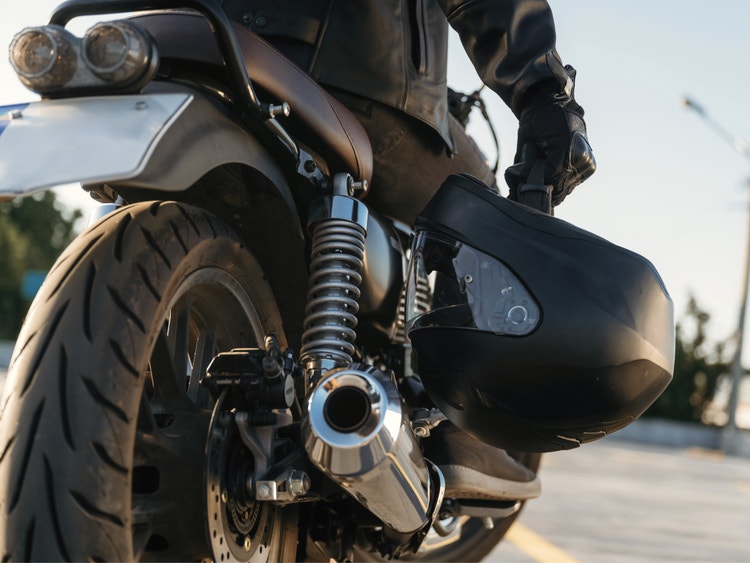 Dettaglio ravvicinato di un motociclista che regge un casco nero accanto alla ruota posteriore della sua moto.