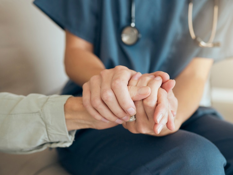 Un operatore sanitario in divisa blu stringe calorosamente la mano di un paziente anziano.