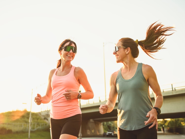 Due donne sorridenti con occhiali da sole corrono all&#39;aperto al tramonto, vestite con canotte sportive.