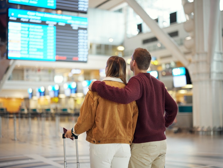 Coppia di spalle con valigia guarda il tabellone dei voli partenze in un grande aeroporto.