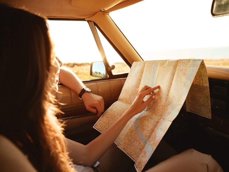 Donna lato passeggero in auto legge e indica un punto su una mappa stradale cartacea aperta.