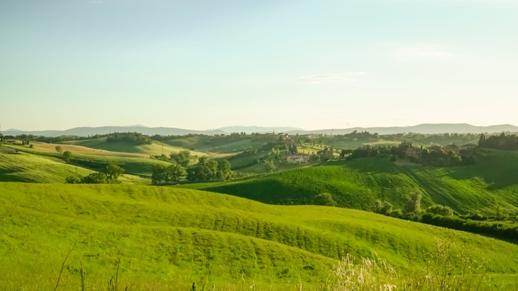 Colline verdi e alberate sotto un cielo chiaro, con piccoli borghi e case rurali in lontananza.