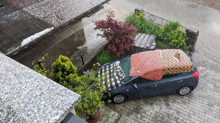 Auto scura parcheggiata sotto una forte grandinata, protetta da coperte colorate stese su tetto, parabrezza e cofano.
