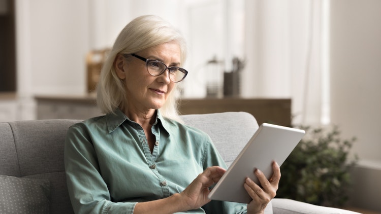 Donna con occhiali seduta sul divano mentre usa un tablet, con sfondo domestico sfocato.